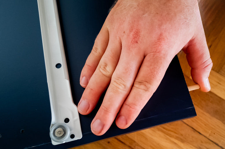 A hand touches a bracket on the underside of a shelf turned over and placed against the surface of a wooden floor.